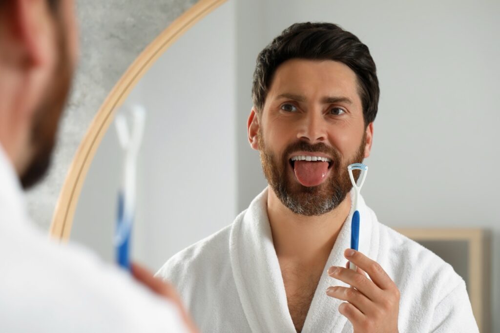 Man looks at his reflection in mirror and sticks out his tongue while holding plastic tongue scraping tool. 