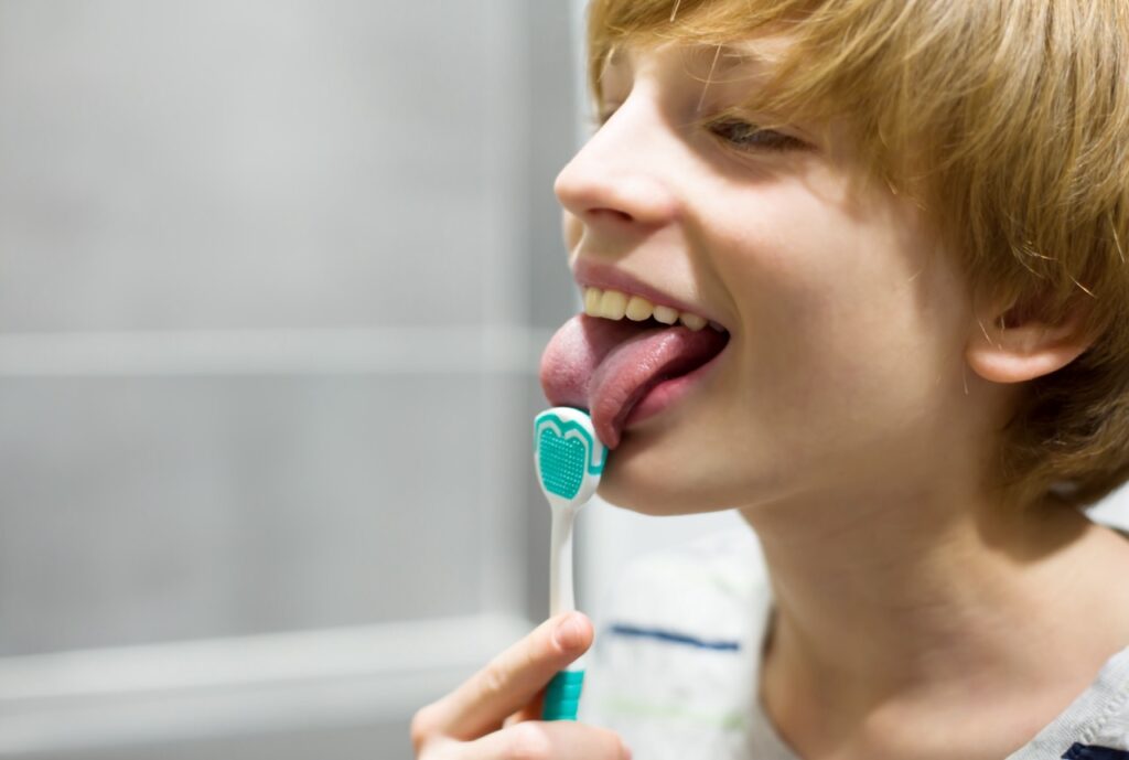 Young adolescent boy smiles as he uses tongue scraping tool to clean his tongue’s surface.
