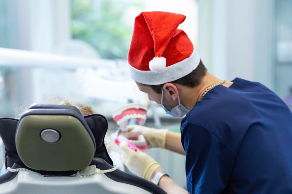 Dentist wearing mask and Santa hat demonstrates proper toothbrushing technique with model teeth to patient in dental chair. 