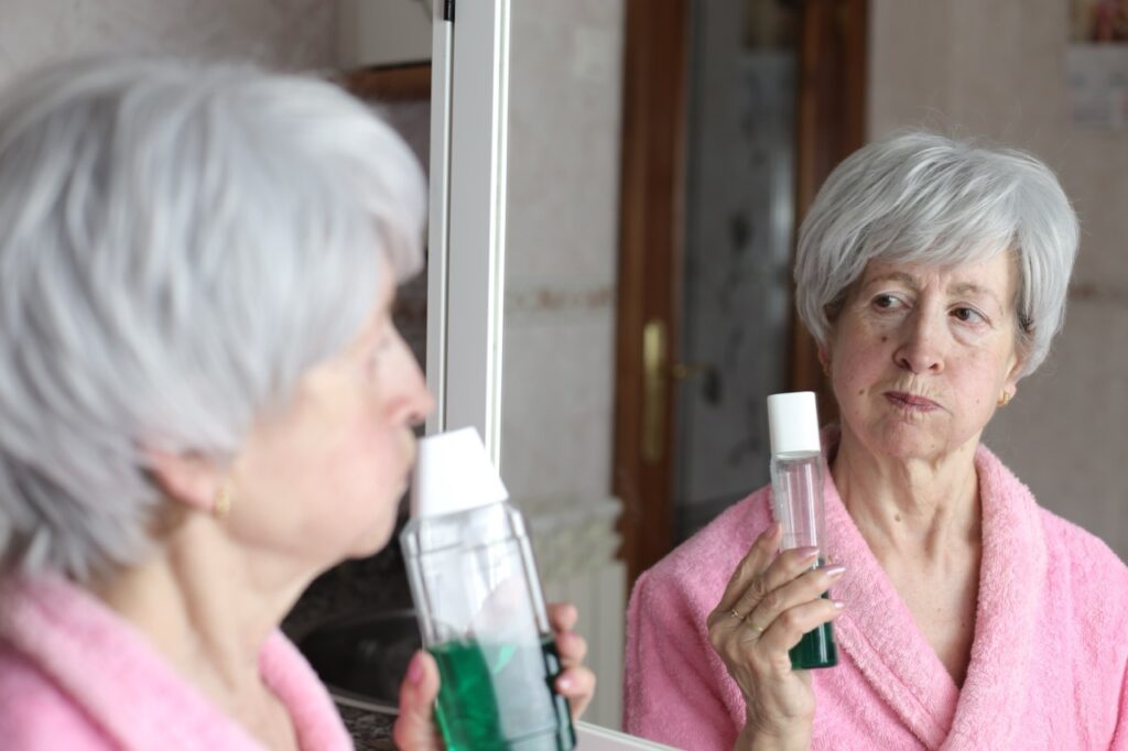 Woman wearing bathrobe stands in front of bathroom mirror, rinsing her mouth with oral microbiome mouthwash.
