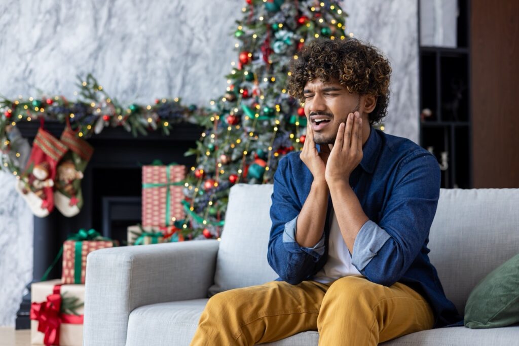 Man sitting on couch in front of decorated Christmas tree winces as he holds hands to jaw, feeling dental pain.