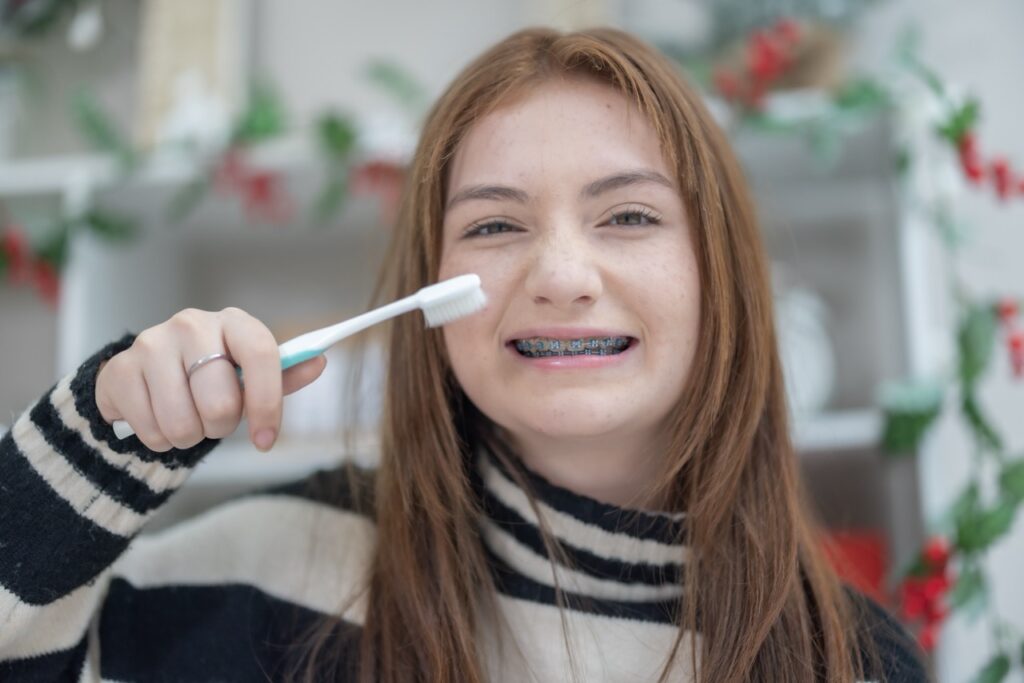 Adolescent girl with braces holds toothbrush and smiles, standing in front of holiday garland. 
