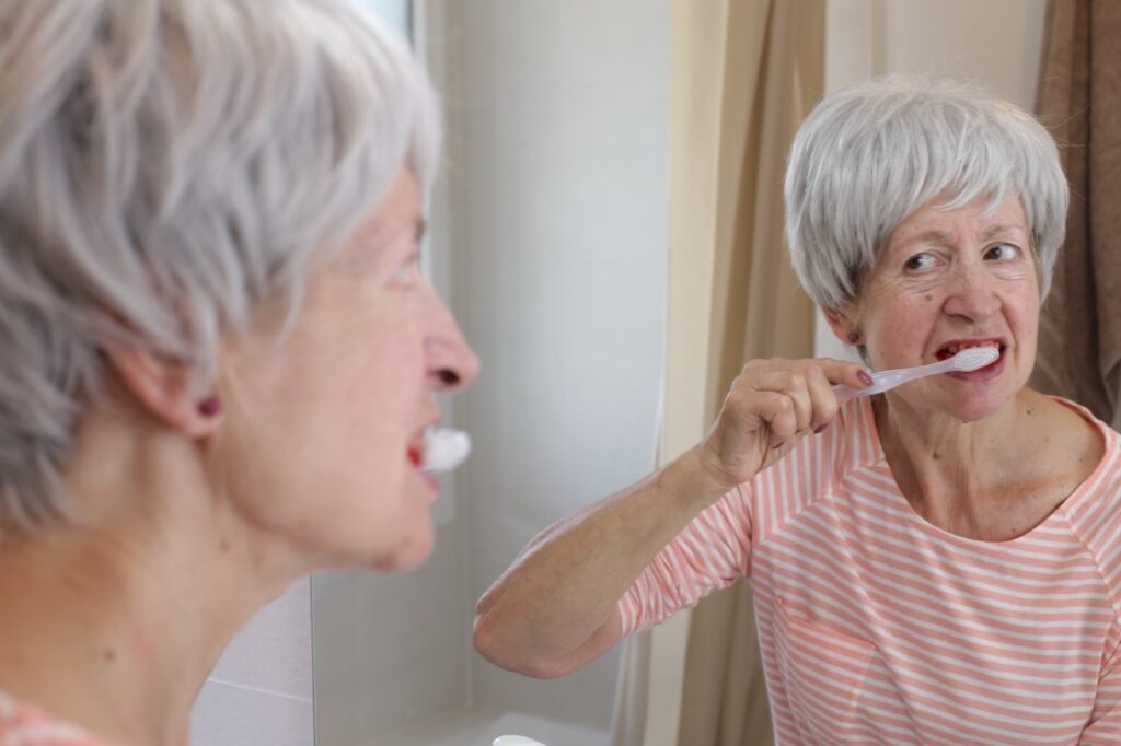Senior woman looks at her reflection in mirror with concern as she feels pain while brushing her teeth, a sign of gum disease.