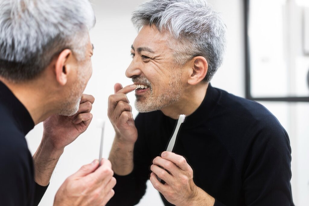 Man stops brushing teeth to look at his inflamed gums in bathroom mirror, a sign of gum disease. 