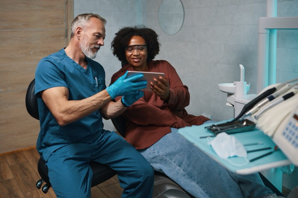 Woman in dental chair reviews dentist’s treatment plan for her periodontal disease and tooth loss on tablet computer as he sits next to her.