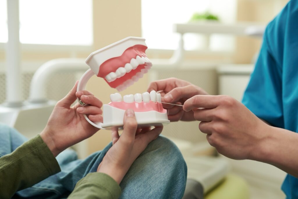 Patient in dental chair holds oversized model of teeth as dentist demonstrates proper flossing technique. 