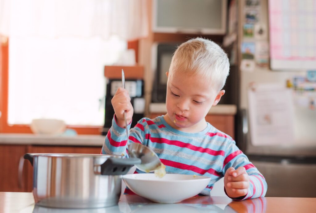 Elementary school-age boy with cleft lip and cleft palate sits at kitchen counter, ladling soup from pot into bowl.
