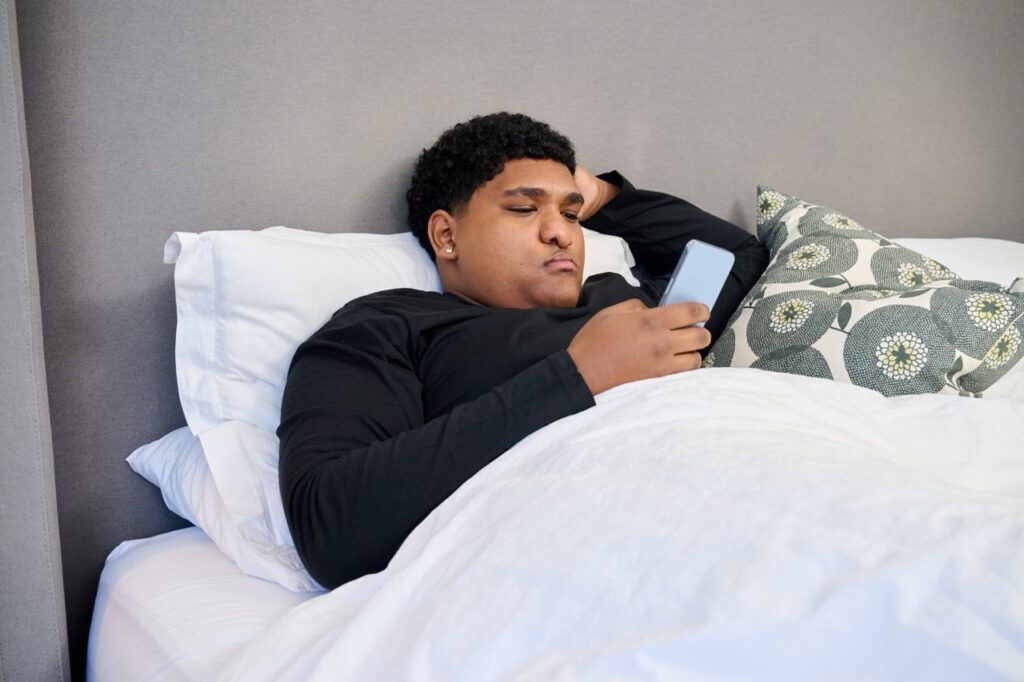 Man with faint scar to upper lip, indicating past cleft palate surgery, lies in bed, scrolling on his smartphone. 
