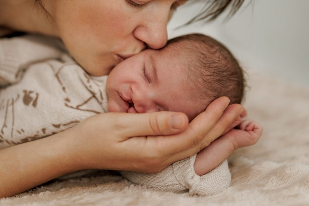 Mother leans over her young infant who has a cleft lip and cleft palate, gently kissing the baby’s right cheek. 