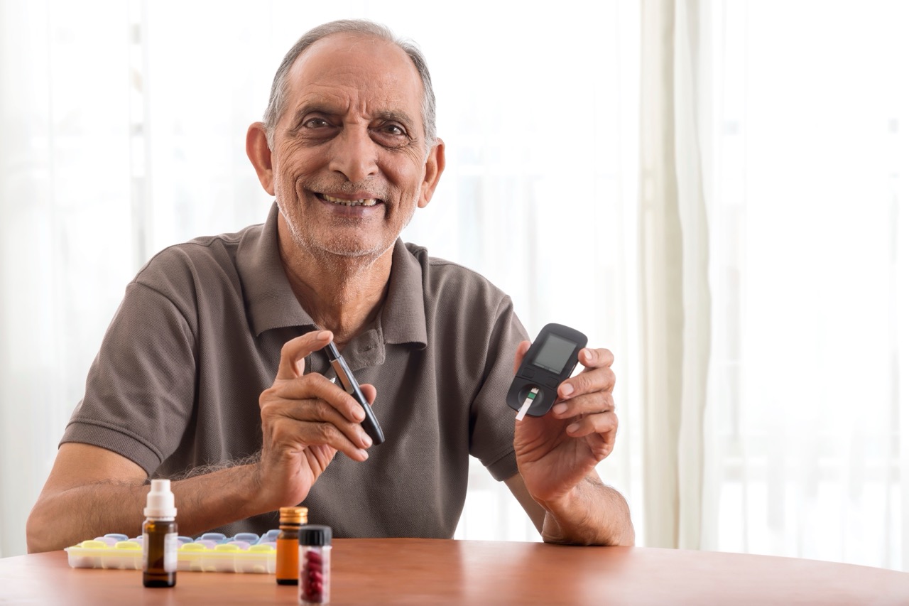 Senior man with diabetes smiles as he sits at dining room table, holding digital glucose level monitor. 