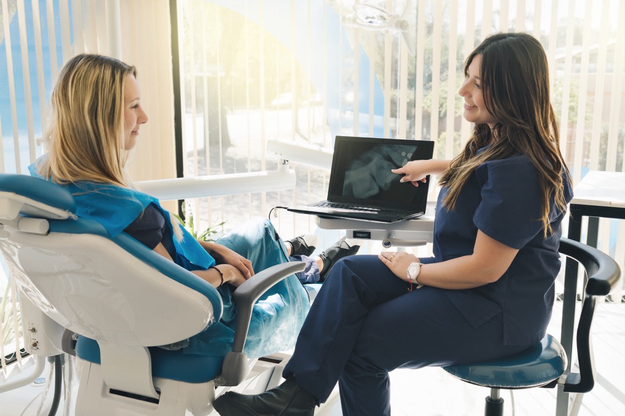 Dentist sits next to woman in dental chair, showing her AI-enhanced imaging of patient’s teeth.