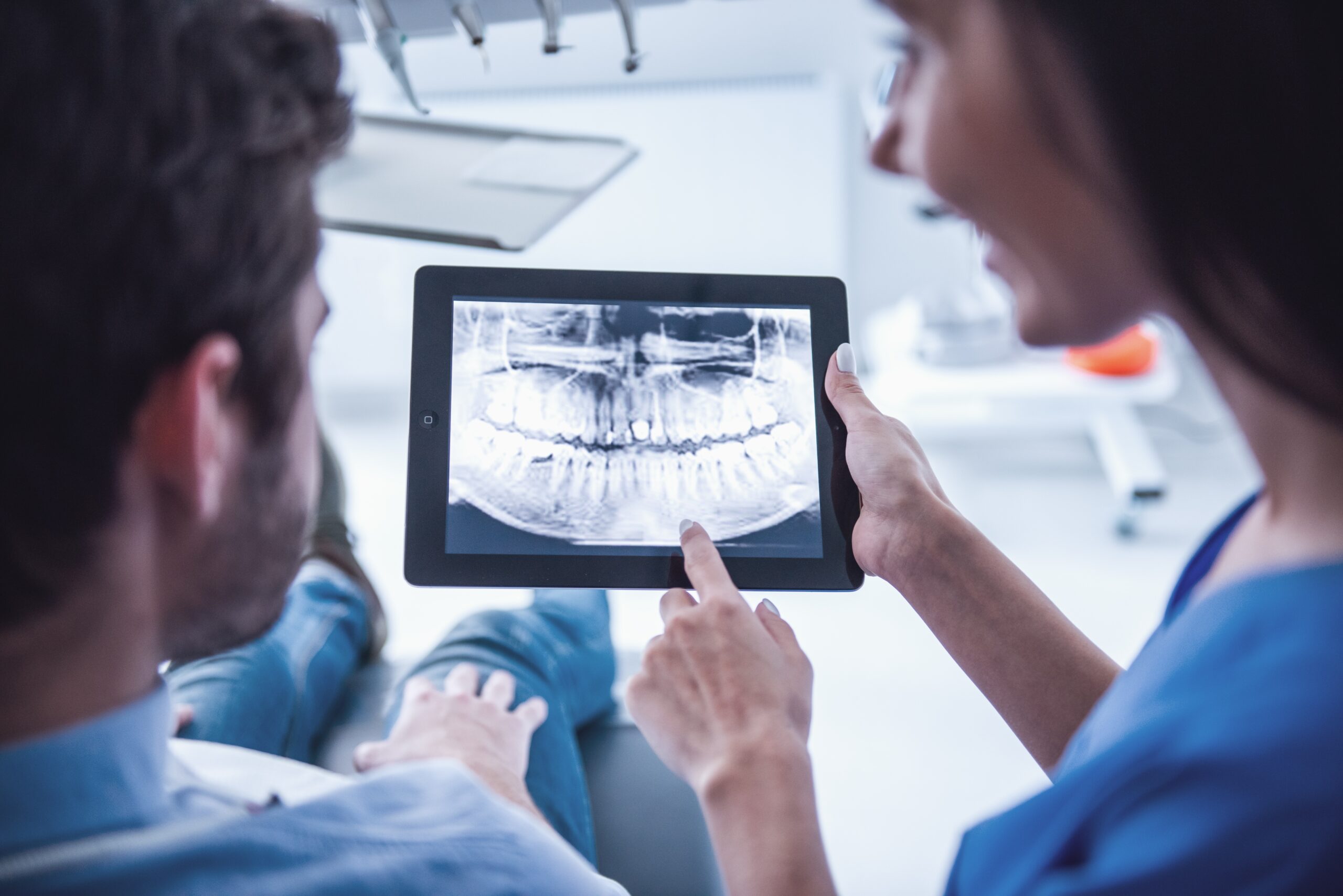 Dentist holds tablet computer to review dentistry digital imaging with her patient, who sits in dental chair.
