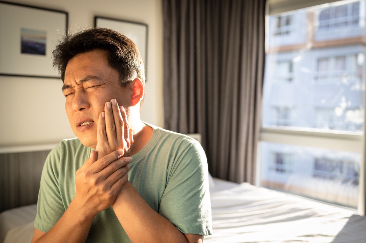 Man sitting on bed winces as he holds his left hand to his left cheek, feeling pain that may mean he needs a root canal.