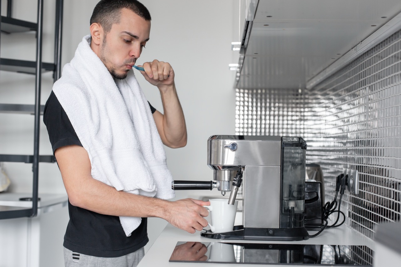 Man stands in his kitchen, brushing his teeth before drinking the coffee he is brewing in a mug on the countertop.