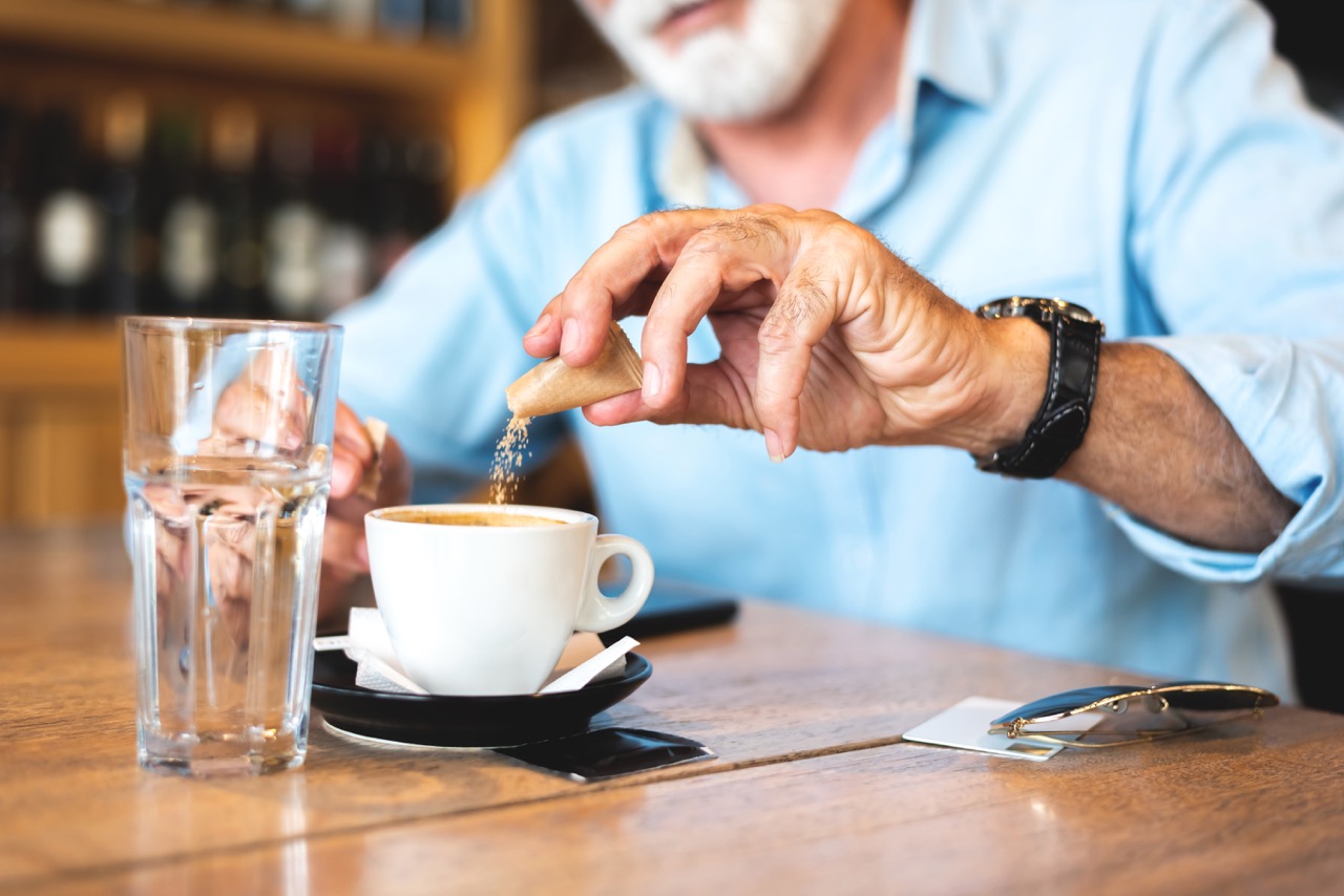 Bearded man sits at table, pouring packet of brown sugar into his morning cup of coffee.