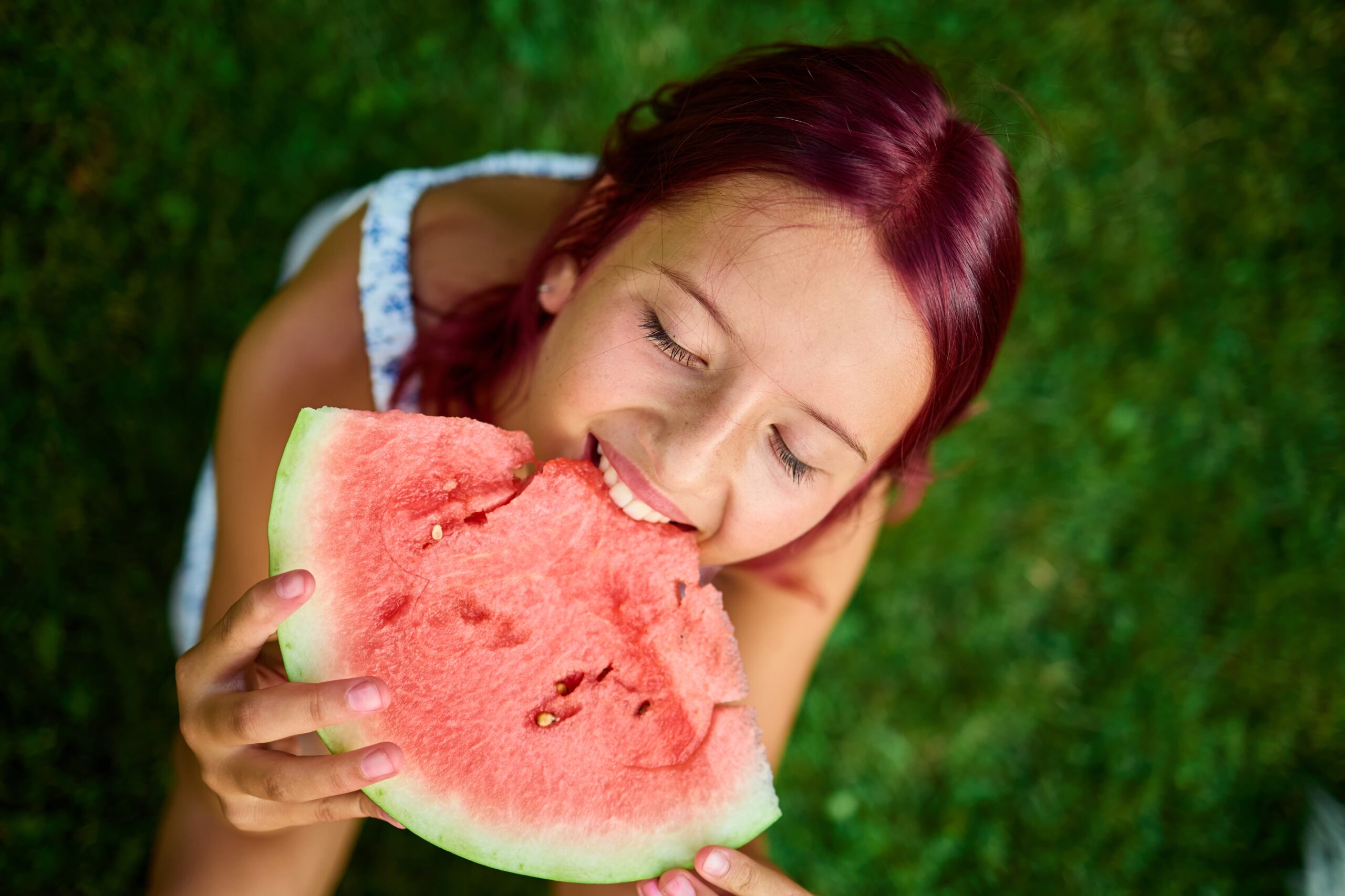 Teenage girl smiles, closes her eyes, and takes a bite out of a watermelon slice with her straight teeth.